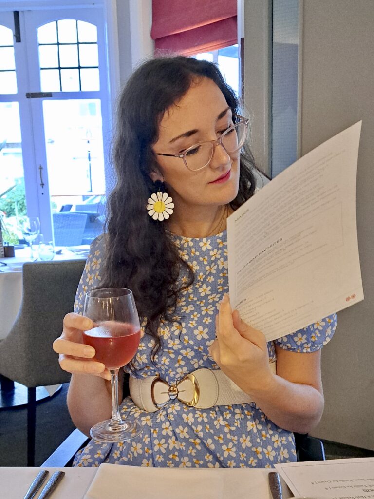 Georgina, a white brunette female, wearing a blue dress holding a menu with a glass of wine